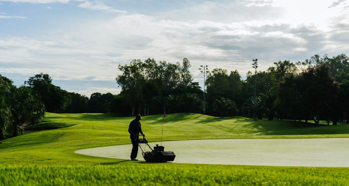 Groundskeeper mowing the green on a golf course during a sunny day, showcasing lush landscapes and maintenance.